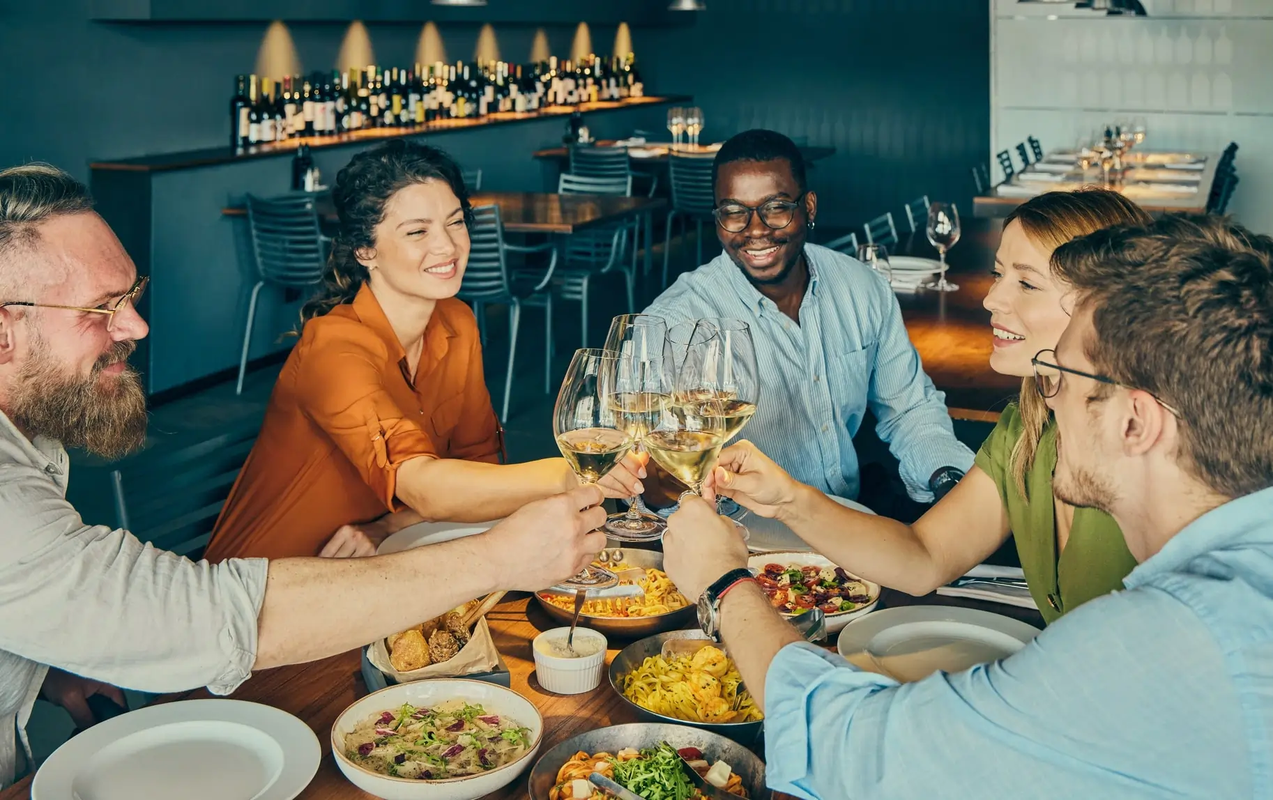 A diverse group of smiling people clinking wine glasses over a dinner table, symbolizing hospitality, connection, and shared dining in a warm restaurant setting.